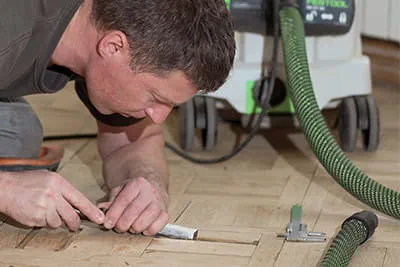 Parketo meistras Jarekas Gawina pasilenkęs virš grindų medžio kaltu preciziškai šalina nelygumus ir pažeidimus